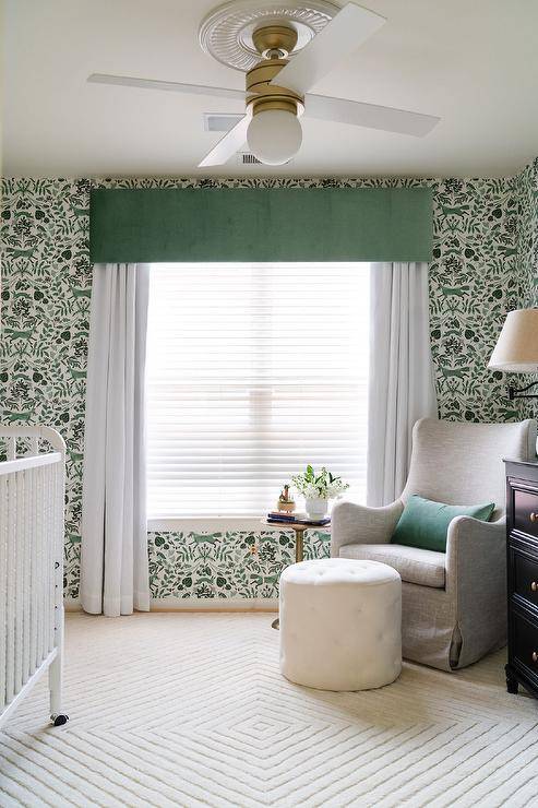 Green and white boy's nursery features a gray wingback glider with a white tufted pouf, a pedestal vocalizing table, a surf rug, a untried valance box over white curtains, untried and white forest wallpaper and a white ceiling fan.