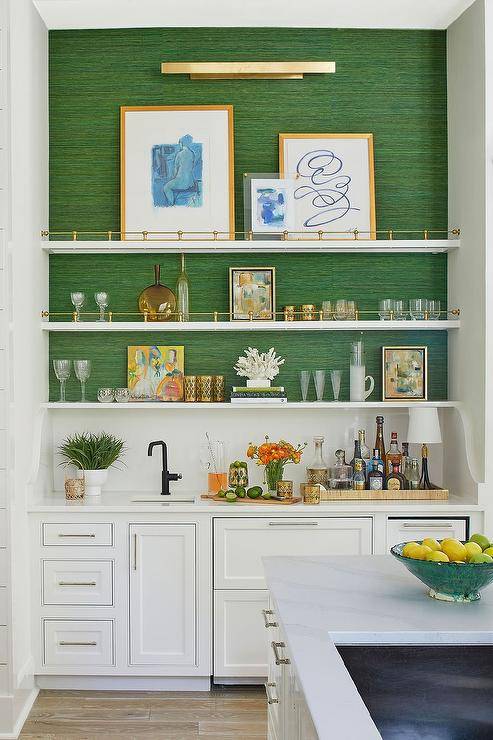 A brass picture light fixed to an emerald green grasscloth wall lights styled and stacked white and gold shelves fixed above white bar cabinets donning polished nickel pulls and finished with a sink with an oil rubbed bronze faucet kit.