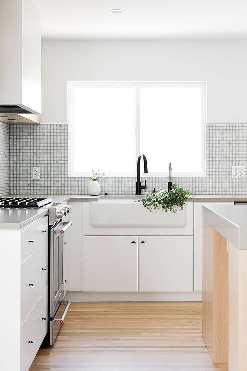 Gray grid backsplash tiles partially frame a window located above a farmhouse sink fitted to white flat front cabinets with oil robbed bronze pulls and paired with an oil rubbed bronze faucet fixed to a gray quartz countertop.