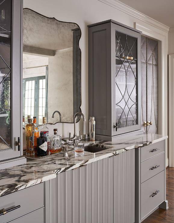 A pink and black marble countertop accents gray wet bar drawers donning black pulls and completed with a small square stainless steel sink and a satin nickel faucet. The faucet is mounted beneath a smoke mirror flanked by seeded glass cabinets.