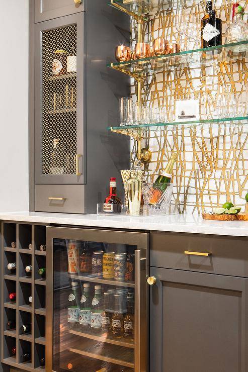 Contemporary wet bar design features gray bar cabinets with brass grille doors and brass pulls, floating glass bar shelves, a glass front fridge and a built in wine rack.