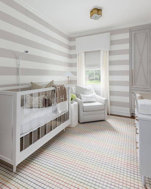 A colorful vintage grid rug sits beneath a wood and lucite crib accented with a brown Hermes throw blanket and placed against a gray striped wall. A white glider is positioned in a corner in front of a brass floor lamp and a window covered in white curtains layered behind a white valance. Gray built in cabinets are stacked and finished with x-fronts.