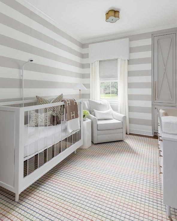 A colorful vintage grid rug sits underneath a wood and lucite crib accented with a brown Hermes throw wrap and placed versus a gray striped wall. A white glider is positioned in a corner in front of a contumely floor lamp and a window covered in white taps layered overdue a white valance. Gray built in cabinets are stacked and finished with x-fronts.