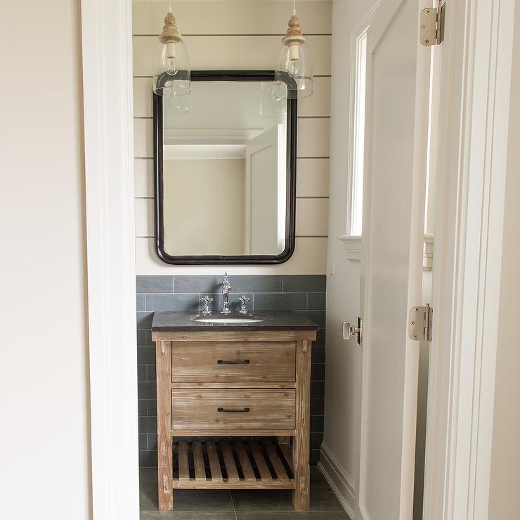 Small white and gray cottage bathroom showcases an eye catching rustic oak single washstand finished with a slated wood shelf, oil rubbed bronze hardware, and a soapstone countertop fitted with an oval undermount sink paired with a polished nickel vintage cross handle faucet. The faucet is positioned in front of gray linear backsplash tiles and beneath a curved black vanity mirror mounted on a white shiplap wall lit by two glass light pendants.