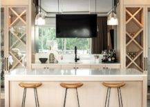 Three backless wood and iron stools are placed at a tan bar island. Behind the island, a TV is mounted in front of a mirrored backsplash over a small round sink with a matte black faucet. The TV is flanked by glass front built-in wine rack cabinets.