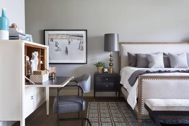 Master bedroom features an ivory French drop down secretary desk, gray leather desk chair, bed with gray and white bedding and a black nightstand lit by a brass and silver lamp.