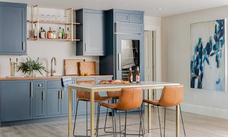 Open concept space with blue kitchenette cabinets and wooden countertops finished with brass hardware and a brass gooseneck faucet sink. A marble and brass bar table with a pub height showcases a set of brown molded leather barstools for an updated and stylish touch.