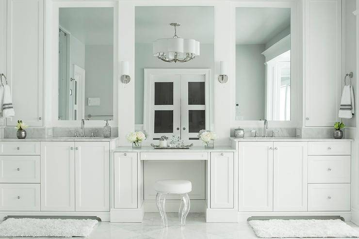 Gorgeous master bathroom boasts his and hers washstands topped with gray marble under framed mirrors flanking a drop down makeup vanity paired with a round white stool with lucite legs illuminated by sconces and a Chandelier.