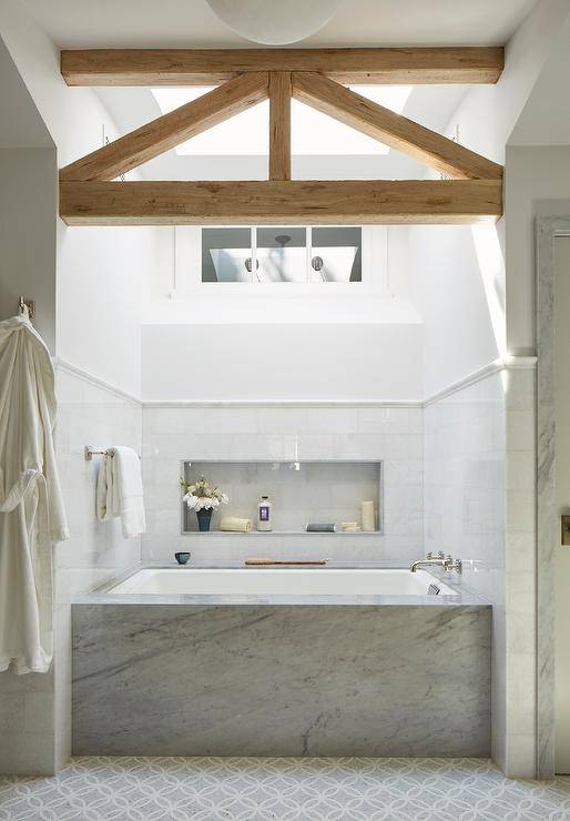 A gorgeous trestle ceiling accents a bathroom boasting a skylight located over a marble clad drop-in bathtub fixed to white and gray marble mosaic floor tiles against a marble tiled wall. A large niche is located about the tub.