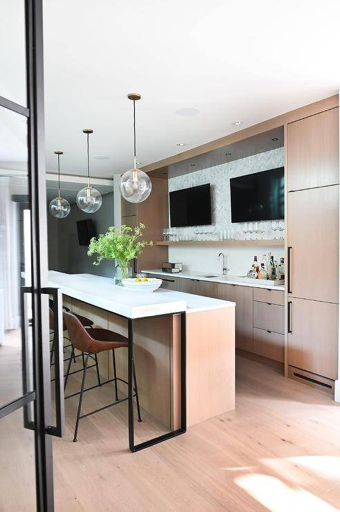 Large modern wet bar design features side-by-side TVs mounted on gray marble chevron backsplash tiles over light brown veneer cabinets. Brown leather stools sit at a quartz topped wet bar under clear globe pendants.