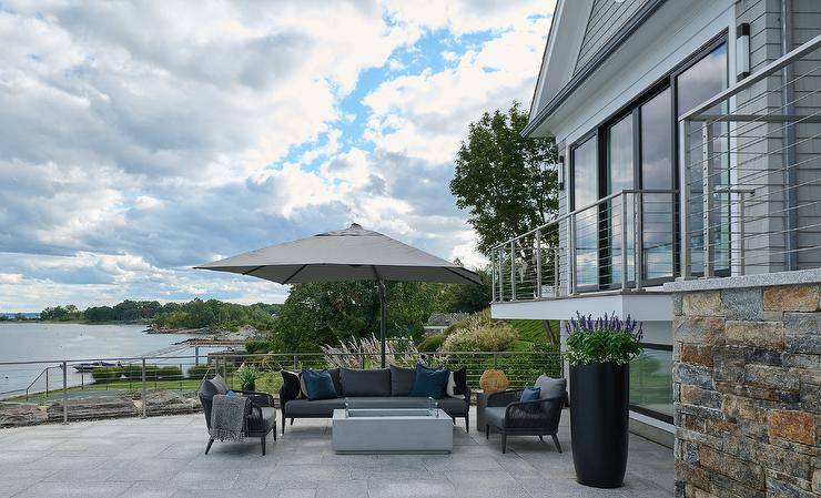 A concrete fire pit warms a black rope sofa and black rope chairs accented with black cushions topped with blue pillows and shaded by a gray outdoor umbrella.