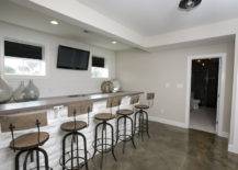Basement features long wet bar lined with industrial barstools across from bar sink under flatscreen TV flanked by windows.