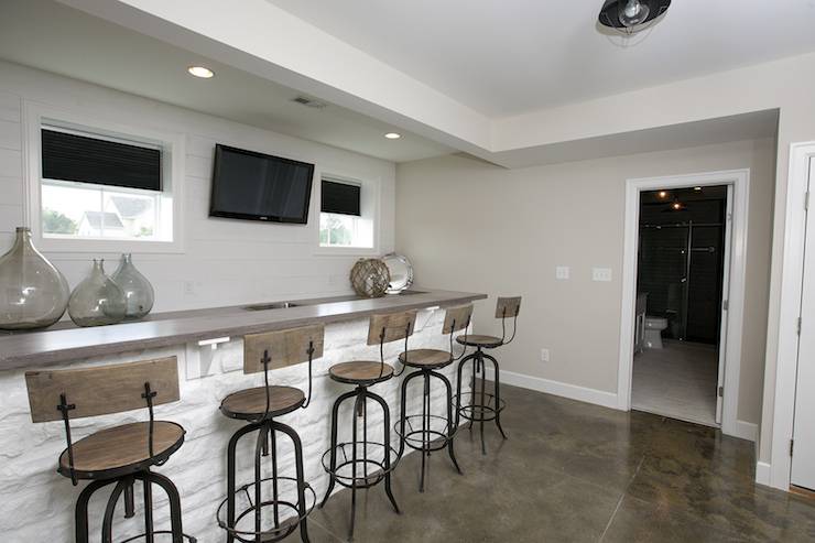 Basement features long wet bar lined with industrial barstools across from bar sink under flatscreen TV flanked by windows.