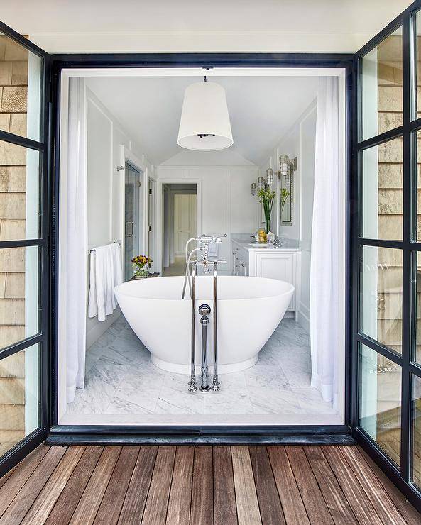 White cottage bathroom features a white tapered drum pendant light which hangs over a freestanding bathtub placed in front of steel doors dressed in white curtains which open to the deck.