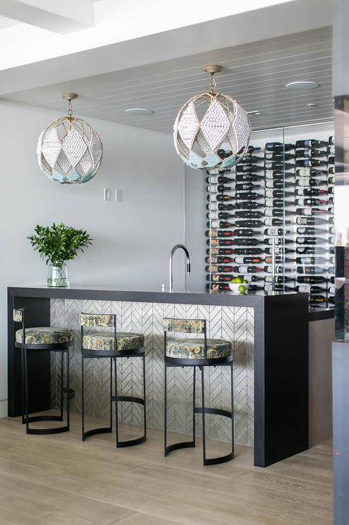 Modern wet bar features yellow and gray barstools at a silver chevron bar island with black quartz waterfall edge illuminated by white beaded sphere chandeliers.