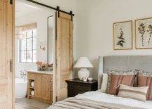 Master bath features salvaged wood doors on rails off a bedroom with a gray bed and tan and white bedding.