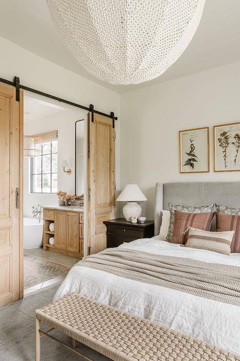 Master bath features salvaged wood doors on rails off a bedroom with a gray bed and tan and white bedding.
