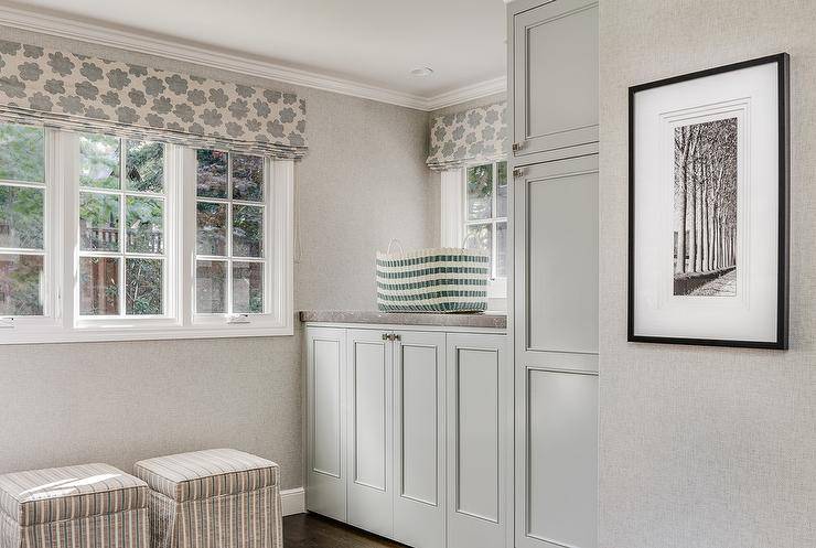 Light gray laundry room features light gray cabinets concealing a washer and dryer topped with zinc lined with a green striped wicker basket. A pair of gray striped skirted ottomans stand under windows dressed in a cream and gray roman shade.