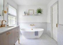 Placed on a marble floor and matched with an antique brass wall mount tub filler, a freestanding bathtub is located in front of a marble wainscot wall lined with a marble chair rail. A styled marble floating shelf is fixed above the bathtub.