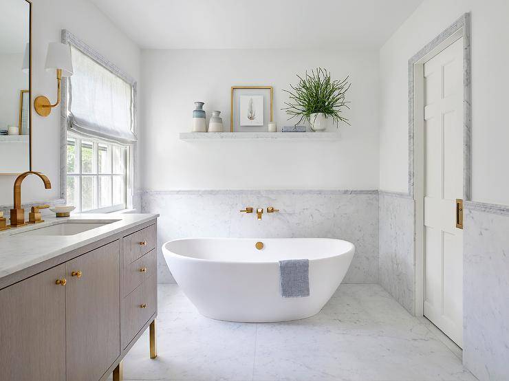 Placed on a marble floor and matched with an antique brass wall mount tub filler, a freestanding bathtub is located in front of a marble wainscot wall lined with a marble chair rail. A styled marble floating shelf is fixed above the bathtub.
