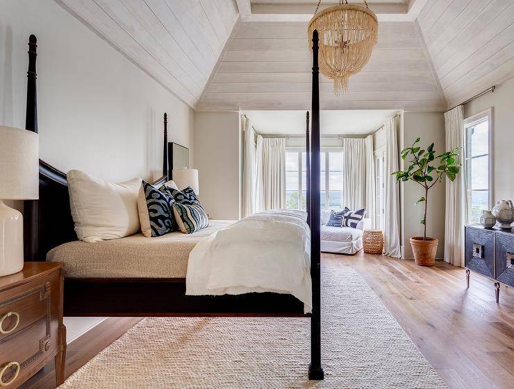 Made Goods Carmen Chandelier elegantly hanging with a tan fringe style from a gray plank vaulted ceiling in a transitional bedroom furnished with a black 4 poster bed, French nightstands and white ceramic lamps.