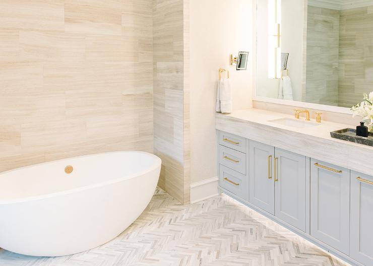 Stunning master bathroom with a light gray washstand and light gray marble countertop displaying a white mirror and brass hardware. under cabinet lighting upgrades this design with a luxurious appeal highlighting white and gray herringbone floor tiles. An oval freestanding tub against a light gray staggered tiled wall invites a spa like look to the bathroom appeal.