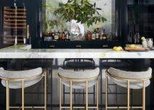 Chic home bar features gold and gray stools placed at a u-shaped glossy blue island topped with a marle countertop. A smoked glass mirror hangs above a glossy blue cabinets located beside a glass front wine fridge.