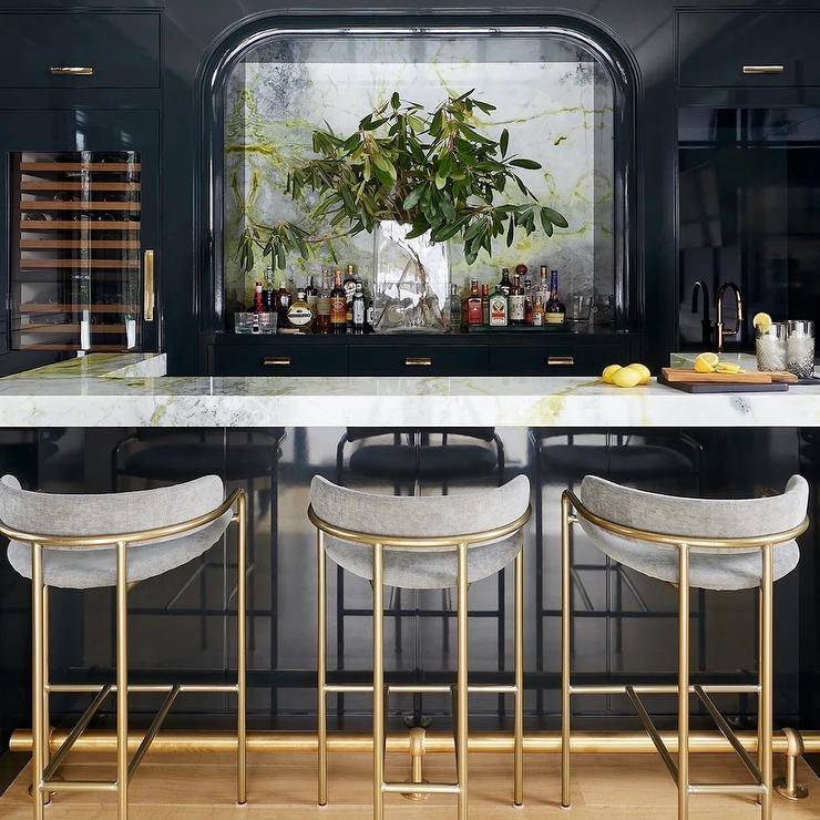 Chic home bar features gold and gray stools placed at a u-shaped glossy blue island topped with a marle countertop. A smoked glass mirror hangs above a glossy blue cabinets located beside a glass front wine fridge.