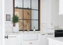 An interior window is located over a farmhouse sink paired with 2 polished nickel faucets and fixed over white cabinets topped with a marble countertop mounted against a marble backsplash.