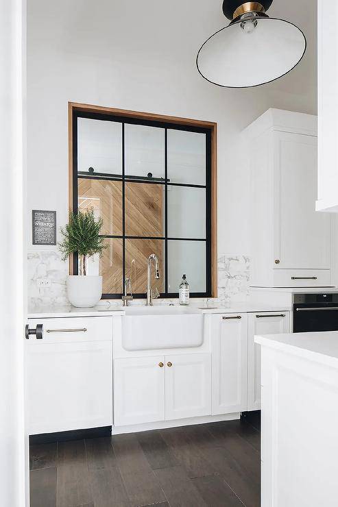 An interior window is located over a farmhouse sink paired with 2 polished nickel faucets and fixed over white cabinets topped with a marble countertop mounted against a marble backsplash.
