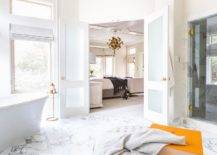 Frosted glass double doors open to an en suite master bathroom boasting a marble tiled floor and a brass cigar table placed beside a catty corner freestanding bathtub.