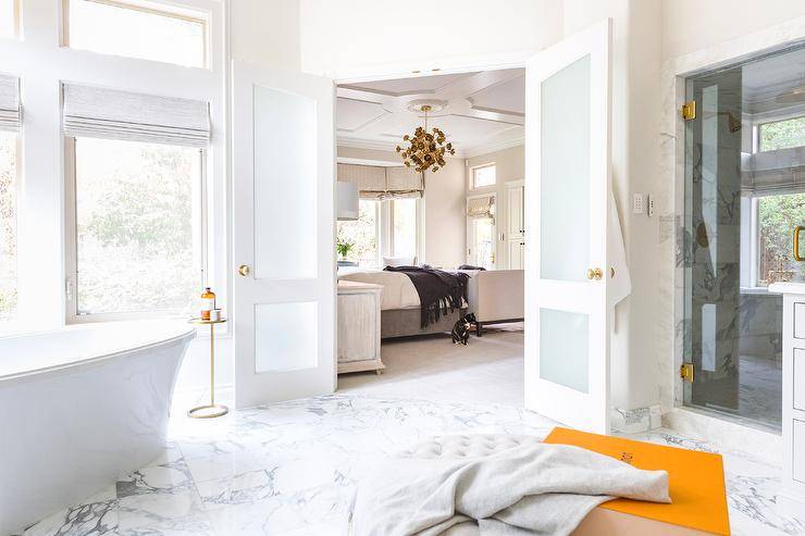 Frosted glass double doors open to an en suite master bathroom boasting a marble tiled floor and a brass cigar table placed beside a catty corner freestanding bathtub.