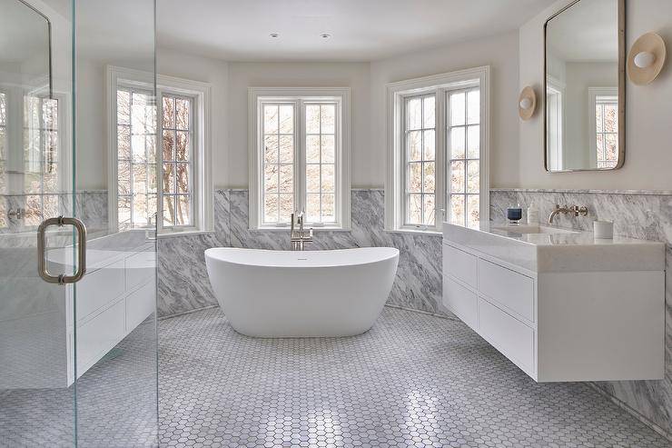 Master bathroom bay windows are partially framed by a gray marble slab backsplash and are positioned over an oval freestanding bathtub with a polsihed nickel floor mount tub filler fixed against marble hexagon floor tiles.