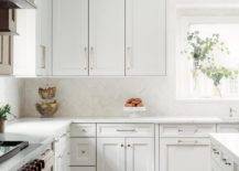 White kitchen cabinets accented with polished nickel hardware are fixed against white herringbone pattern backsplash tiles and beneath white upper cabinets lined with white crown moldings.