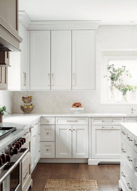 White kitchen cabinets accented with polished nickel hardware are fixed against white herringbone pattern backsplash tiles and beneath white upper cabinets lined with white crown moldings.