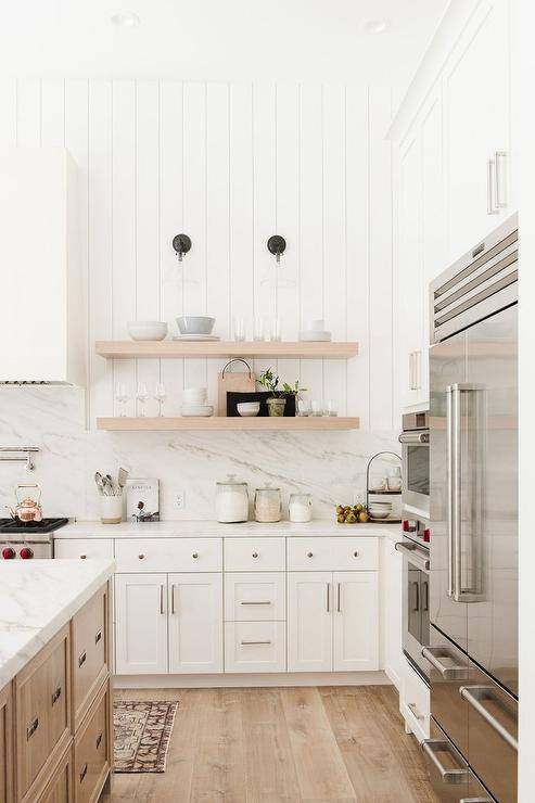 Vertical shiplap walls in a transitional kitchen above white marble backsplash and white quartz countertops on white shaker cabinets. Blond wood floating shelves stack under black and glass sconces for a farmhouse finish.