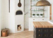 Farmhouse kitchen designed with a corner white brick pizza oven beside brass and glass shelves mounted on a white and gray marble slab backsplash wall. A black apothecary island features a butcher block countertop illuminated by a large aged brass dome pendant light.