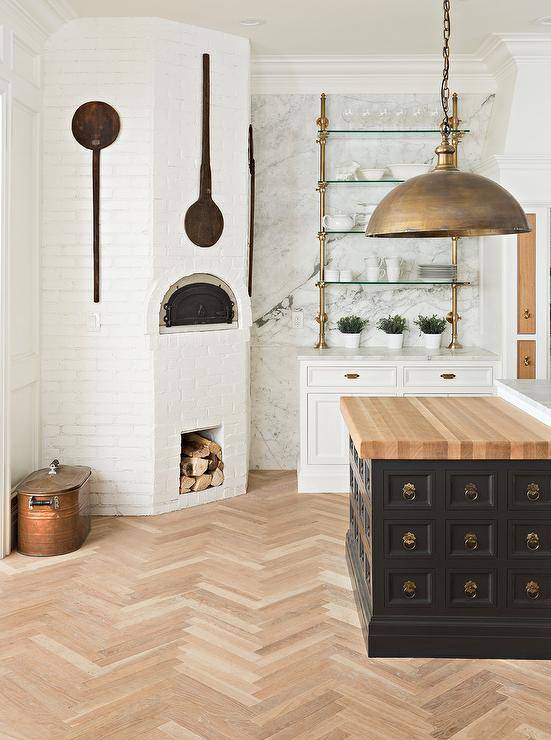 Farmhouse kitchen designed with a corner white brick pizza oven beside brass and glass shelves mounted on a white and gray marble slab backsplash wall. A black apothecary island features a butcher block countertop illuminated by a large aged brass dome pendant light.