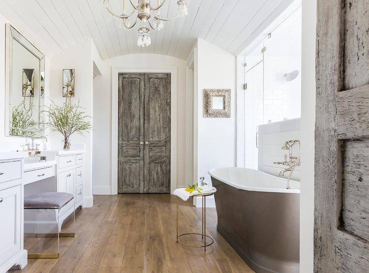 Antique bi-fold doors open to a master bathroom boasting a modern gray cast iron bathtub placed against a shiplap shower wall and lit by a chandelier hung from a white plank barrel ceiling.