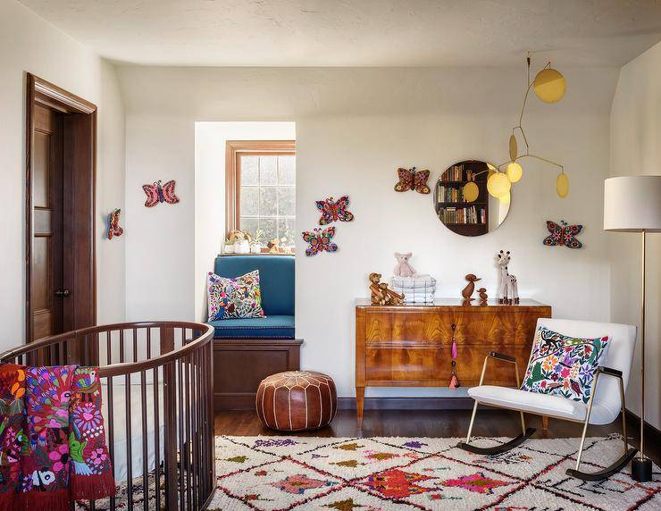 Vintage nursery features a burl wood dresser with pink tassel pulls, an oval wooden crib with colorful blanket, a white vintage nursery rocker accented with an Otomi pillow illuminated by a brass floor lamp, a mini window seat with blue cushion and a colorful rug.