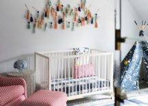 Eclectic red and blue boy's nursery features a white wooden crib placed beneath tassel art hung from a white wall lined with black crown moldings framing a white and gray wallpapered ceiling. The crib is flanked by a red rocker and ottoman and a yellow and blue teepee.