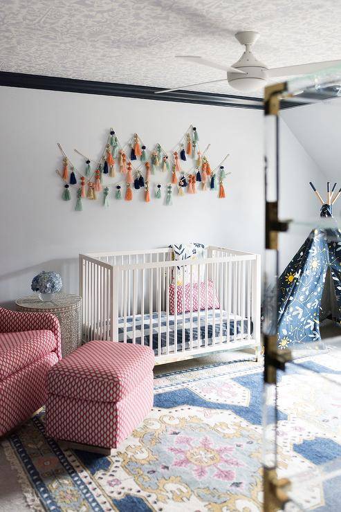 Eclectic red and undecorous boy's nursery features a white wooden crib placed underneath tassel art hung from a white wall lined with woebegone crown moldings framing a white and gray wallpapered ceiling. The crib is flanked by a red rocker and ottoman and a yellow and undecorous teepee.