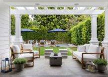 Under a pergola fitted with columns, a rectangular concrete fire pit sits between a brown teak sofa with white cushions and brown teak chairs. The space is completed with gray stripe cube stools.