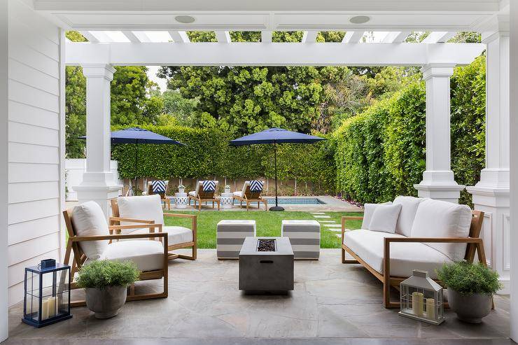 Under a pergola fitted with columns, a rectangular concrete fire pit sits between a brown teak sofa with white cushions and brown teak chairs. The space is completed with gray stripe cube stools.