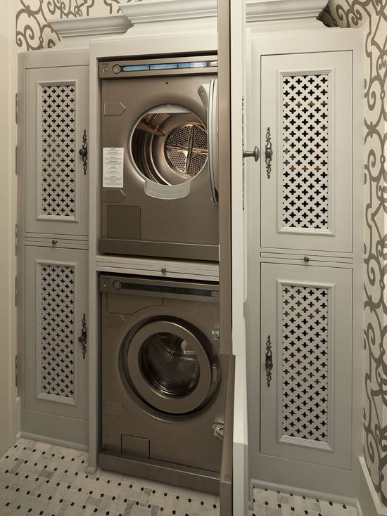 Amazing laundry room with gray scroll trellis wallpaper, Schumacher Manor Gate Wallpaper in Charcoal, and marble basketweave tile laundry room floor. Stacked silver front-load washer and dryer hidden behind gray cabinet doors flanked by gray clover inset panels.