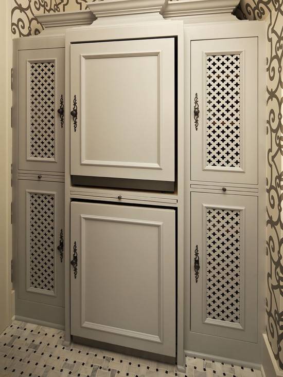 Amazing laundry room with gray scroll trellis wallpaper, Schumacher Manor Gate Wallpaper in Charcoal, and marble basketweave tile laundry room floor. Stacked silver front-load washer and dryer hidden behind gray cabinet doors flanked by gray clover inset panels.