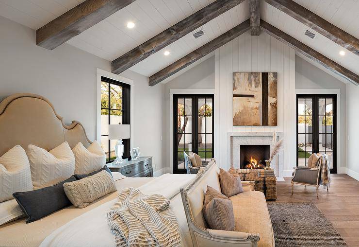 Vertical shiplap fireplace wall in a master bedroom furnished with elegant gray and wood-carved chairs uniting with a woven coffee table decorated with stacked books.