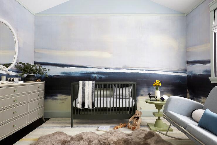 Contemporary blue and gray boy's nursery features black nursery crib, a gray French dresser, a light gray sofa rocker with green avocado accent table, a brown cowhide rug and a blue and gray wall mural.
