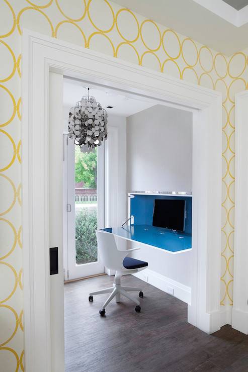White and yellow circle patterned wallpaper frames pocket doors which open to reveal a home office featuring a wall mounted drop down desk with lacquered blue interior lined with a modern white swivel chair below a silver disc pendant hung in front of French doors which lead to the patio.