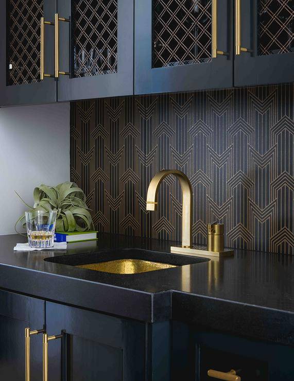 Black wet bar designed with a gold and black backsplash, black quartz countertops, and black cabinets with long brass pulls. A hammered sink adds warmth to the wet bar design finished with black grill cabinet doors.